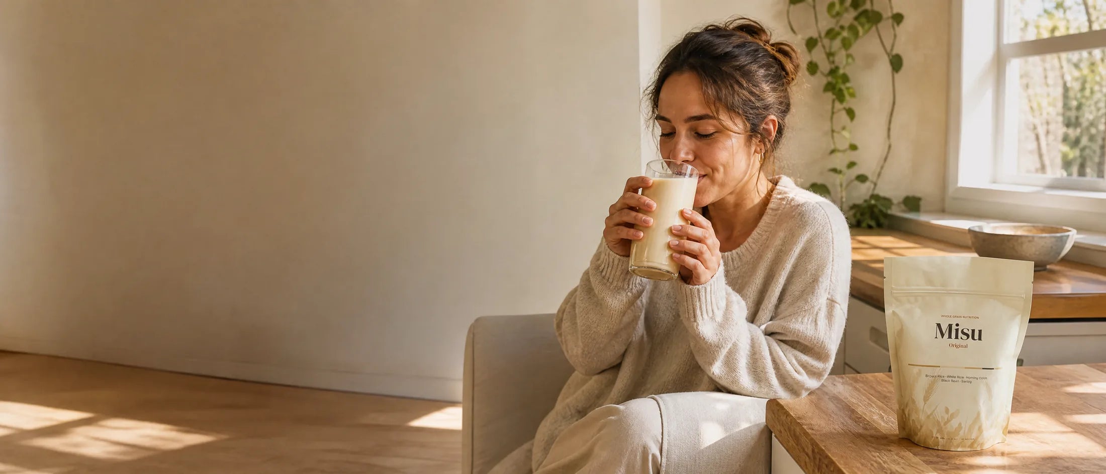 A woman drinking a tall glass of Misu at a sunlit kitchen counter, with the Misu pouch beside her